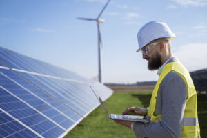 Un professionnel en casque et gilet de sécurité travaille sur un ordinateur portable dans une centrale solaire, éolienne en arrière-plan